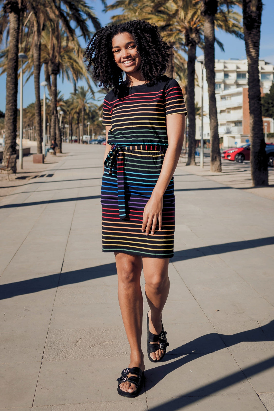 A model wearing a short-sleeved black mini dress with a colorful striped pattern and pockets, standing on a street with palm trees in the background.