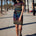 A model wearing a short-sleeved black mini dress with a colorful striped pattern and pockets, standing on a street with palm trees in the background.