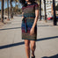 A model wearing a short-sleeved black mini dress with a colorful striped pattern and pockets, standing on a street with palm trees in the background.