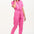 A model wearing a vibrant pink tailored boilersuit with straight legs, half sleeve shirt upper, and an exposed front zip. The suit features contrast cherry embroidery on the bust pocket and is styled with side seam pockets and a tie belt.