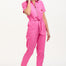 A model wearing a vibrant pink tailored boilersuit with straight legs, half sleeve shirt upper, and an exposed front zip. The suit features contrast cherry embroidery on the bust pocket and is styled with side seam pockets and a tie belt.