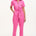 A model wearing a vibrant pink tailored boilersuit with straight legs, half sleeve shirt upper, and an exposed front zip. The suit features contrast cherry embroidery on the bust pocket and is styled with side seam pockets and a tie belt.