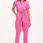 A model wearing a vibrant pink tailored boilersuit with straight legs, half sleeve shirt upper, and an exposed front zip. The suit features contrast cherry embroidery on the bust pocket and is styled with side seam pockets and a tie belt.