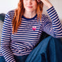 Sugarhill Brighton model wearing a blue and white striped shirt with a pink heart brooch, sitting indoors.