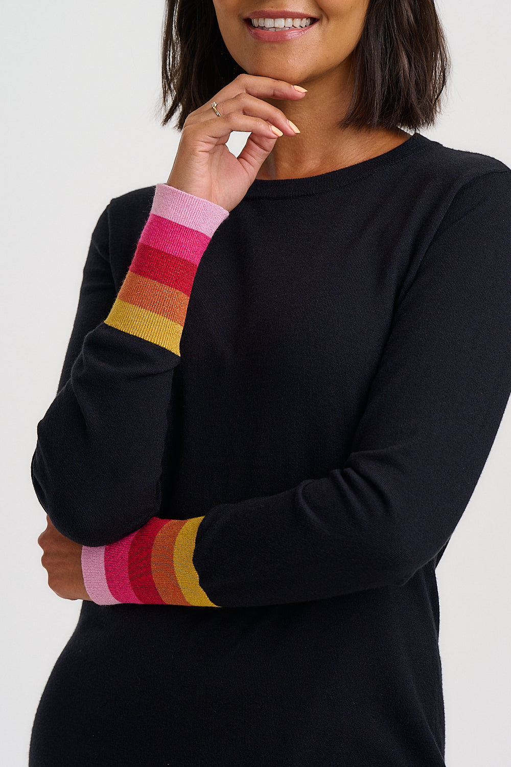 A woman wearing a black long-sleeve knit top with colorful striped ribbed cuffs in pink, orange, red, and yellow, shown against a light gray studio background.