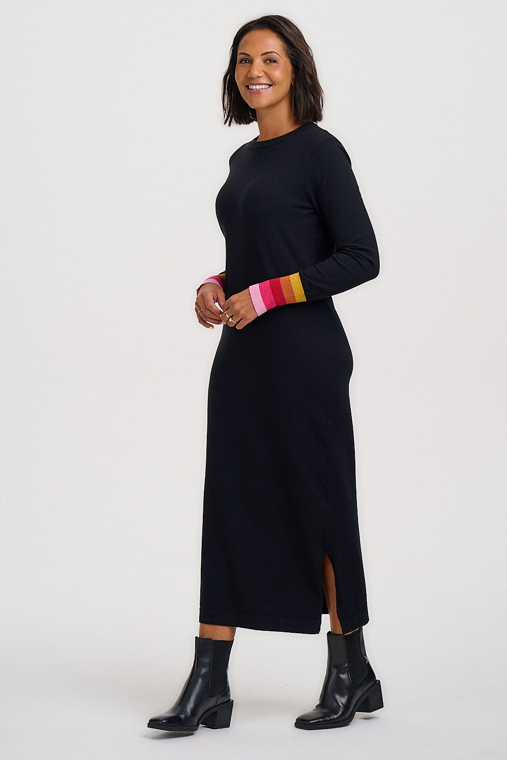 A woman modeling a long-sleeve black midi dress with a colorful striped cuff in pink, orange, yellow, and white, paired with black ankle boots, against a plain white studio background.