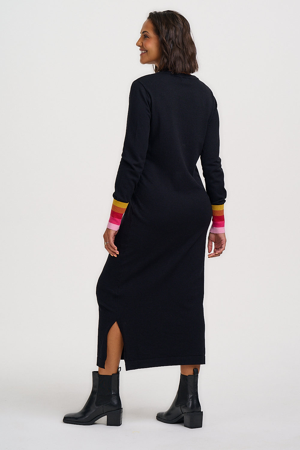 A woman wearing a black long-sleeve midi dress with a side slit and colorful striped cuffs, shown from the back against a plain white studio background.