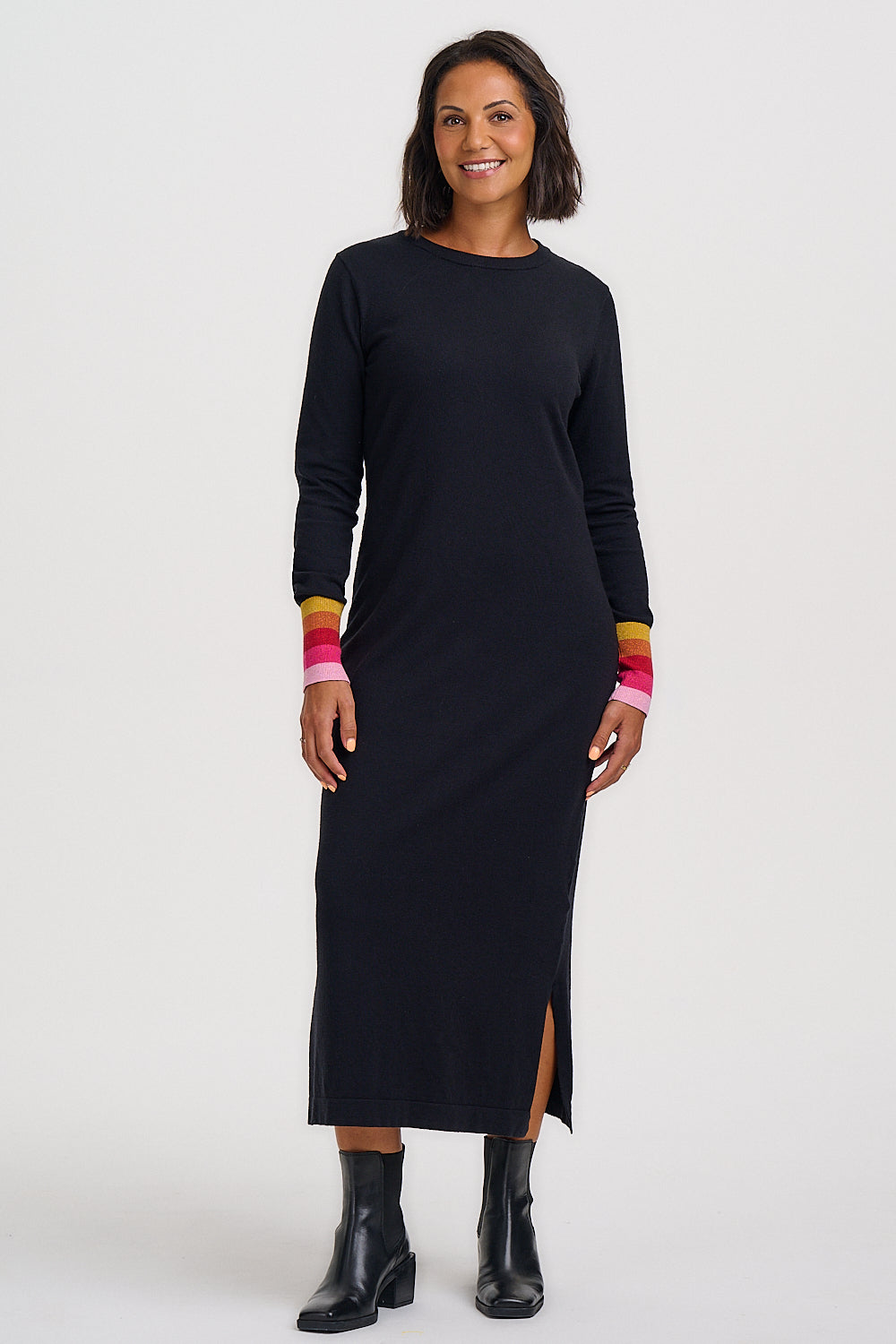 A woman modeling a long-sleeve black midi dress with colorful striped cuffs in pink, orange, and yellow, paired with black ankle boots against a plain light gray studio background.