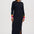 A woman modeling a long-sleeve black midi dress with colorful striped cuffs in pink, orange, and yellow, paired with black ankle boots against a plain light gray studio background.