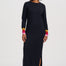 A woman modeling a long-sleeve black midi dress with colorful striped cuffs in pink, orange, and yellow, paired with black ankle boots against a plain light gray studio background.