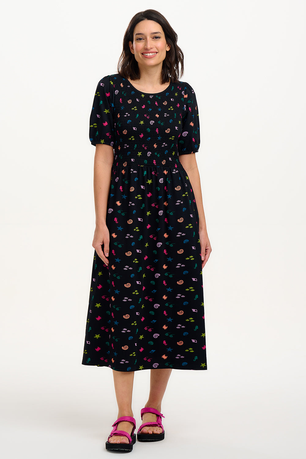 A model wearing a vintage-inspired, black midi dress with a multicolored underwater theme, including small shells and fish on a contrasting black base. The dress features short puff sleeves and a full mini skirt.