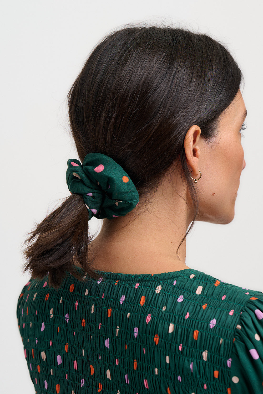 Woman with a green scrunchie in her hair wearing a green top with colorful polka dots on a white background