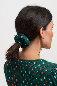 Woman with a green scrunchie in her hair wearing a green top with colorful polka dots on a white background