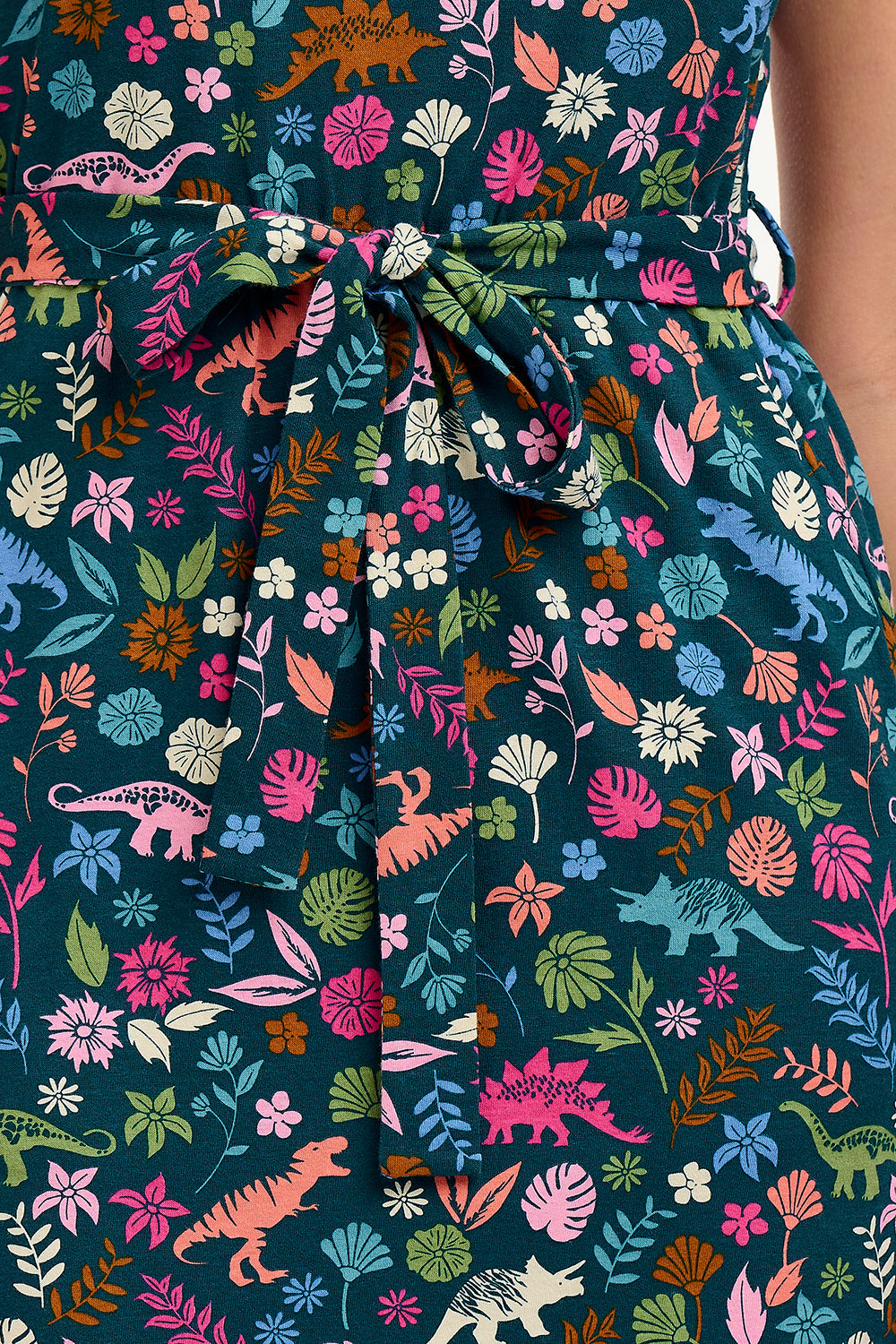 A model wearing a short-sleeved V-neck jersey mini dress with a floral and dinosaur print in various colors on a teal background.