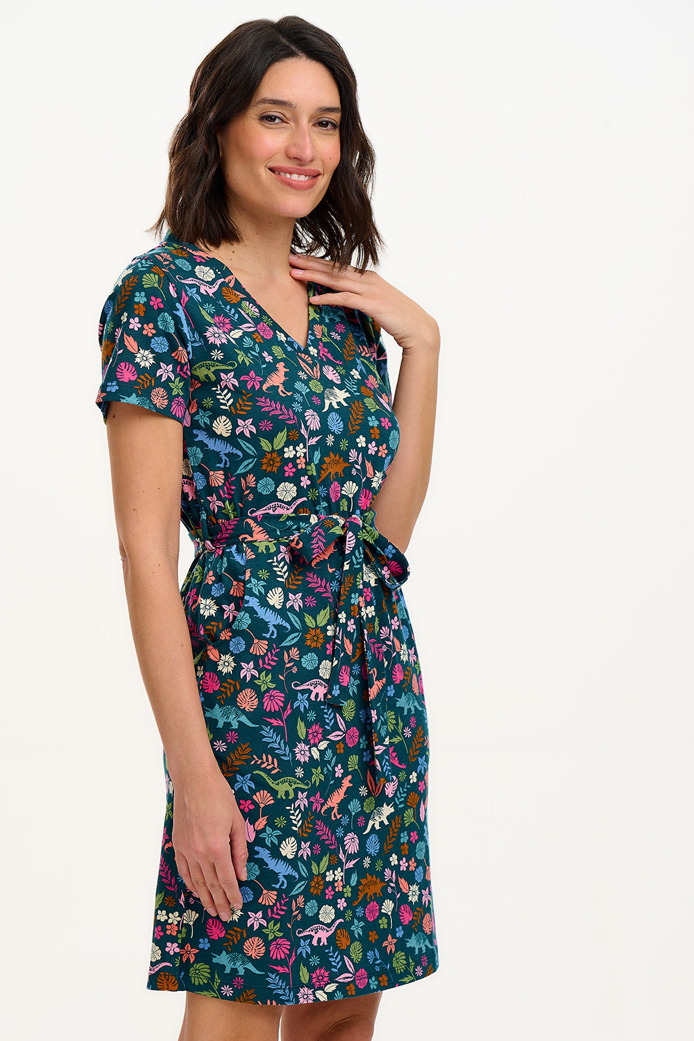 A model wearing a short-sleeved V-neck jersey mini dress with a floral and dinosaur print in various colors on a teal background.