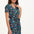 A model wearing a short-sleeved V-neck jersey mini dress with a floral and dinosaur print in various colors on a teal background.