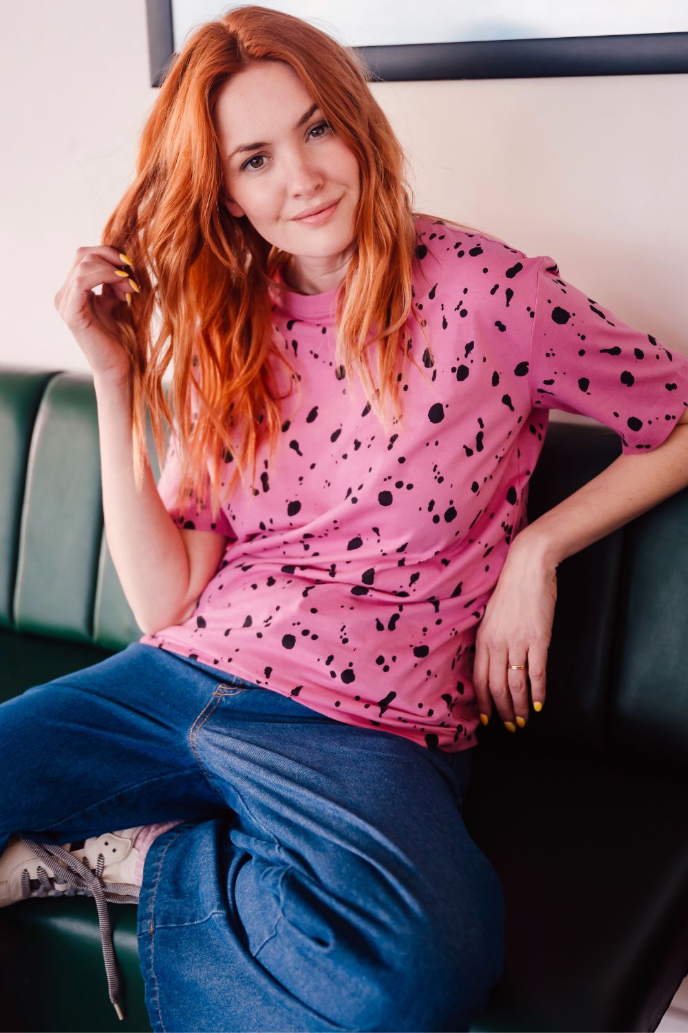 Person with red hair wearing a pink polka dot shirt and blue jeans sitting on a green couch.