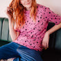 Person with red hair wearing a pink polka dot shirt and blue jeans sitting on a green couch.