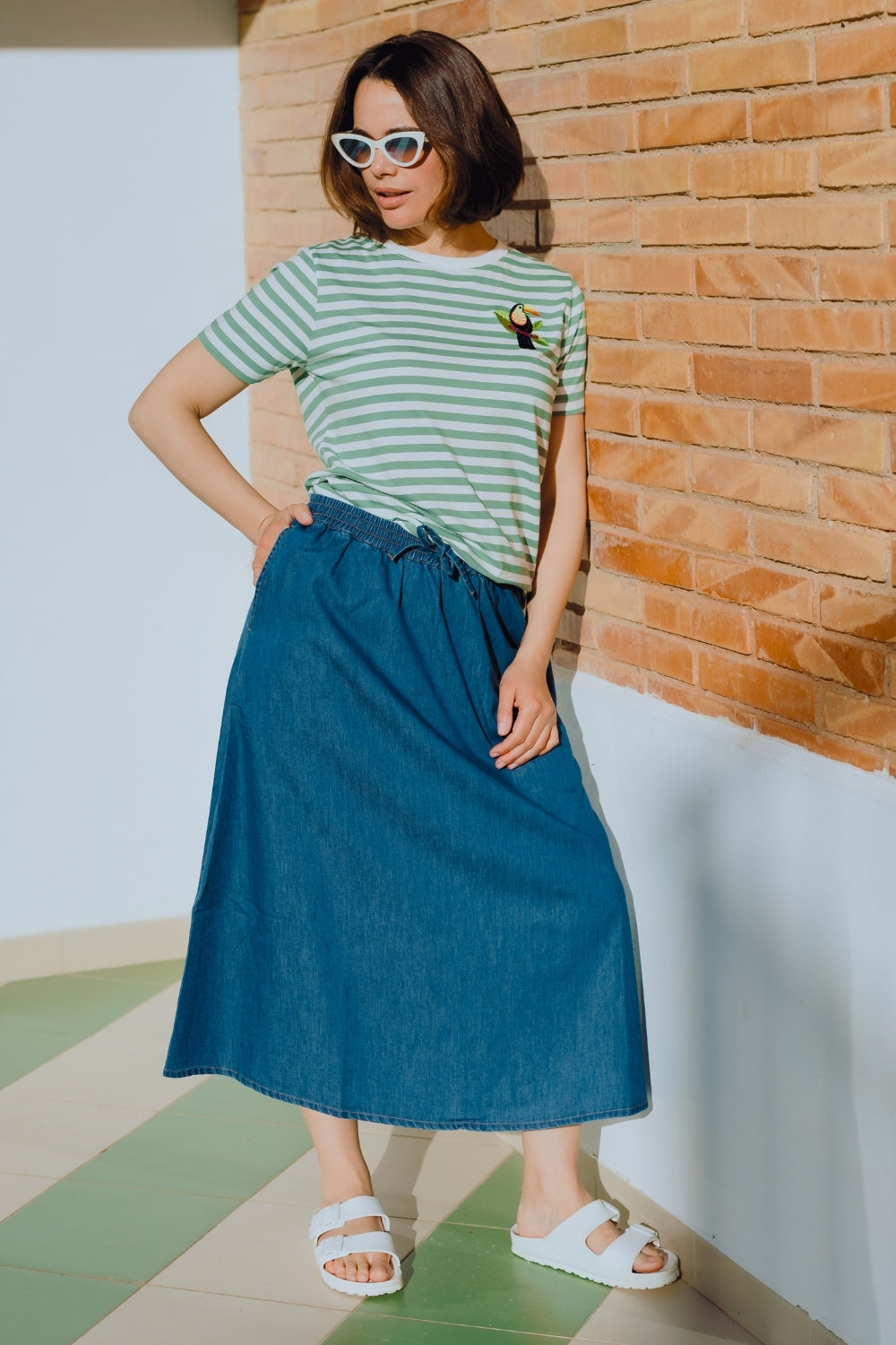 Woman wearing a striped shirt and blue skirt standing against a brick wall.