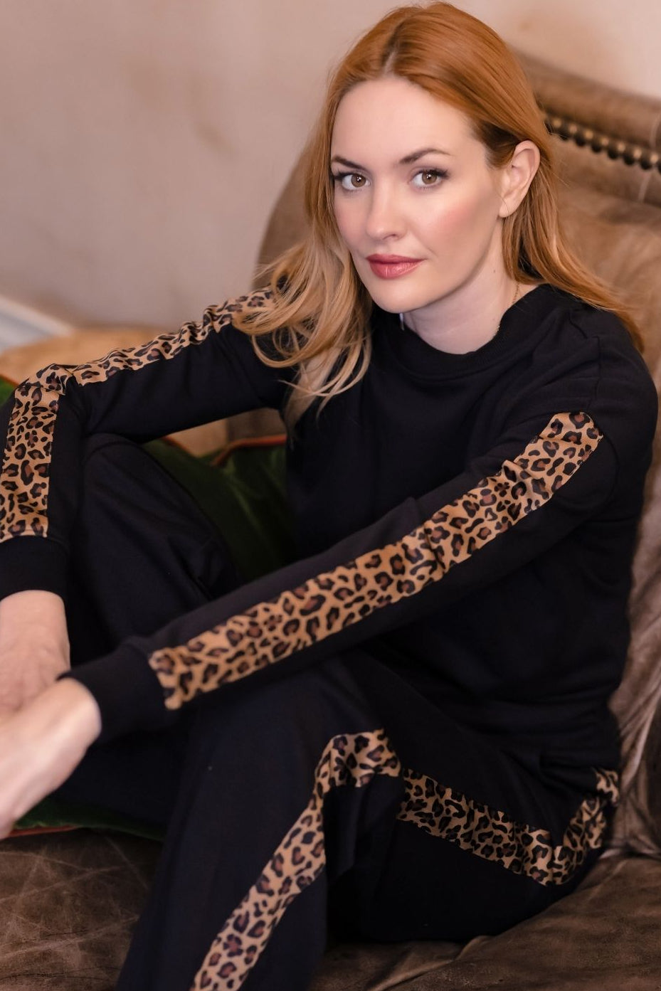A woman with red hair sits on a brown leather couch wearing a black long-sleeve top and matching pants with leopard print stripes on the sleeves and legs.