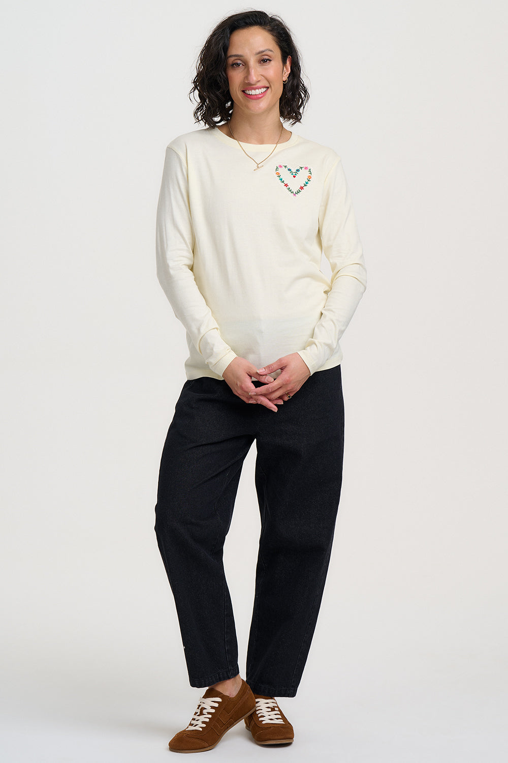 Woman wearing a cream sweater with a heart design, black pants, and brown shoes on a white background