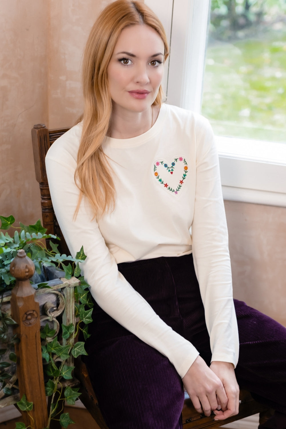 A woman with blonde hair sits on a wooden chair wearing a cream long-sleeve crewneck top with a colorful floral heart on the chest, paired with dark purple pants.