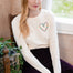A woman with blonde hair sits on a wooden chair wearing a cream long-sleeve crewneck top with a colorful floral heart on the chest, paired with dark purple pants.