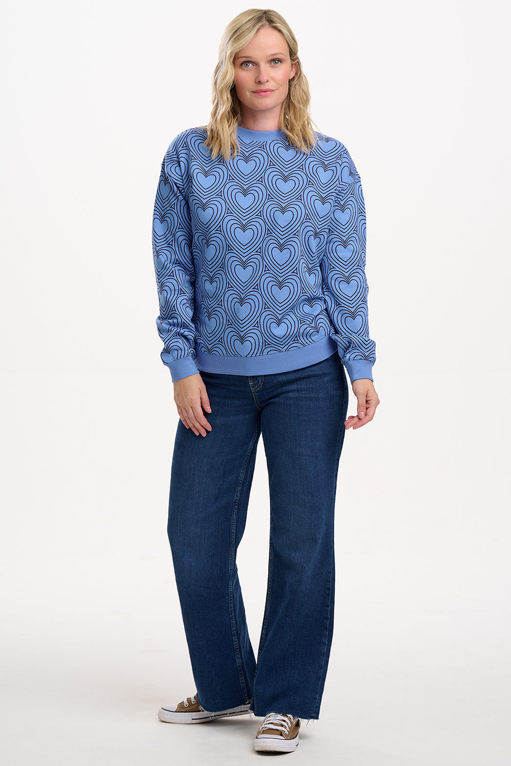 Sugarhill Brighton model wearing a blue sweater with heart pattern and blue jeans on a white background
