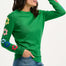 Sugarhill Brighton model wearing a green jumper with flower motifs on the sleeves, paired with blue jeans.
