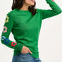 Sugarhill Brighton model wearing a green jumper with flower motifs on the sleeves, paired with blue jeans.