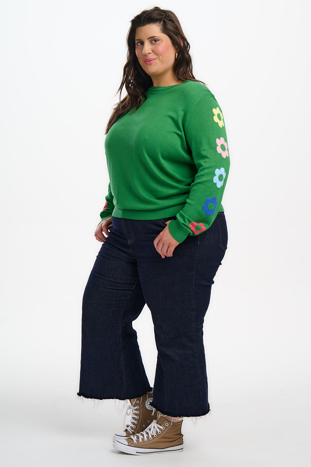 Person wearing a green sweater with colorful flower patterns on sleeves and dark pants against a white background