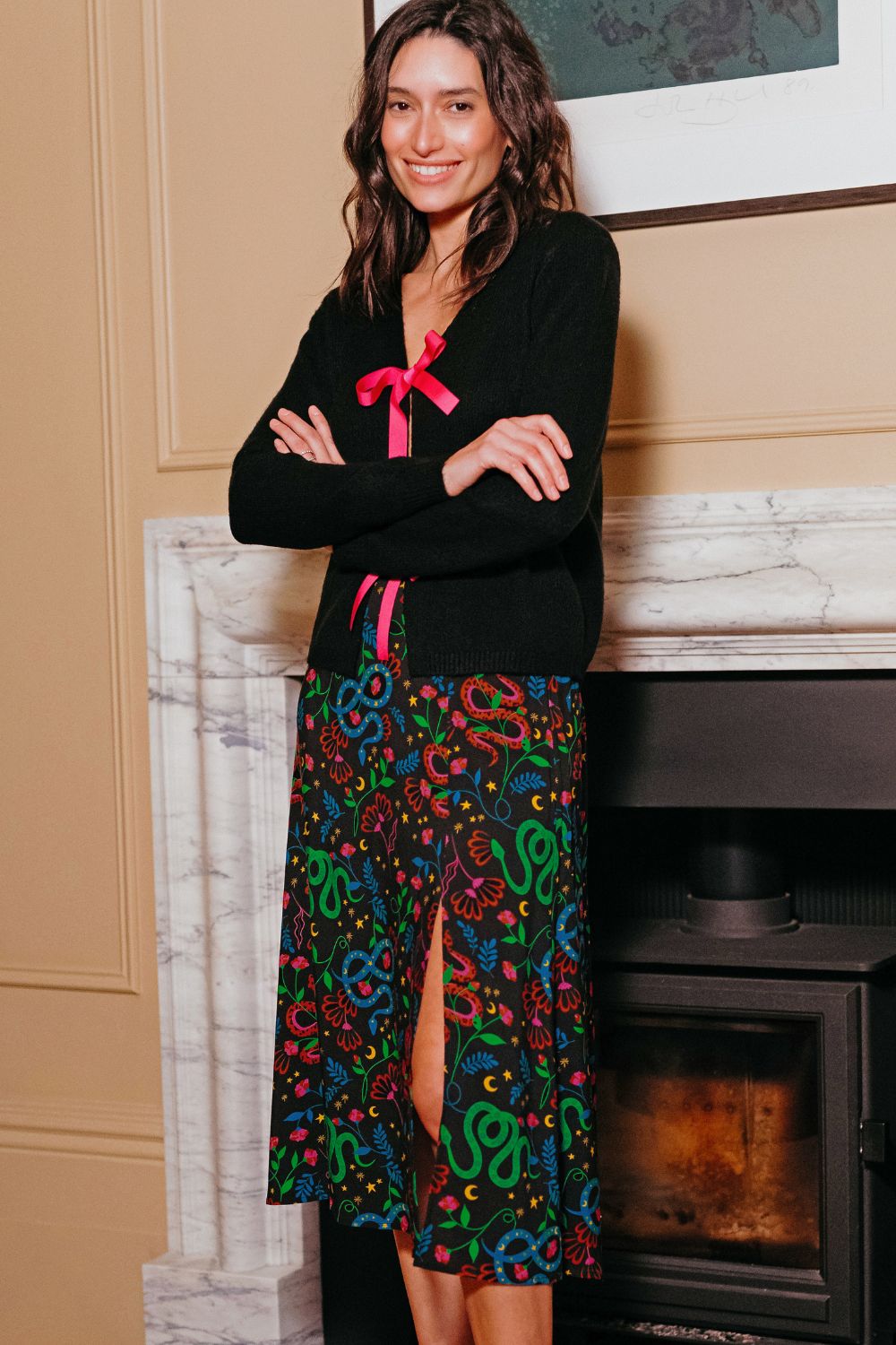 Woman wearing a black top with a pink bow and a colorful snake-patterned skirt standing in front of a fireplace.