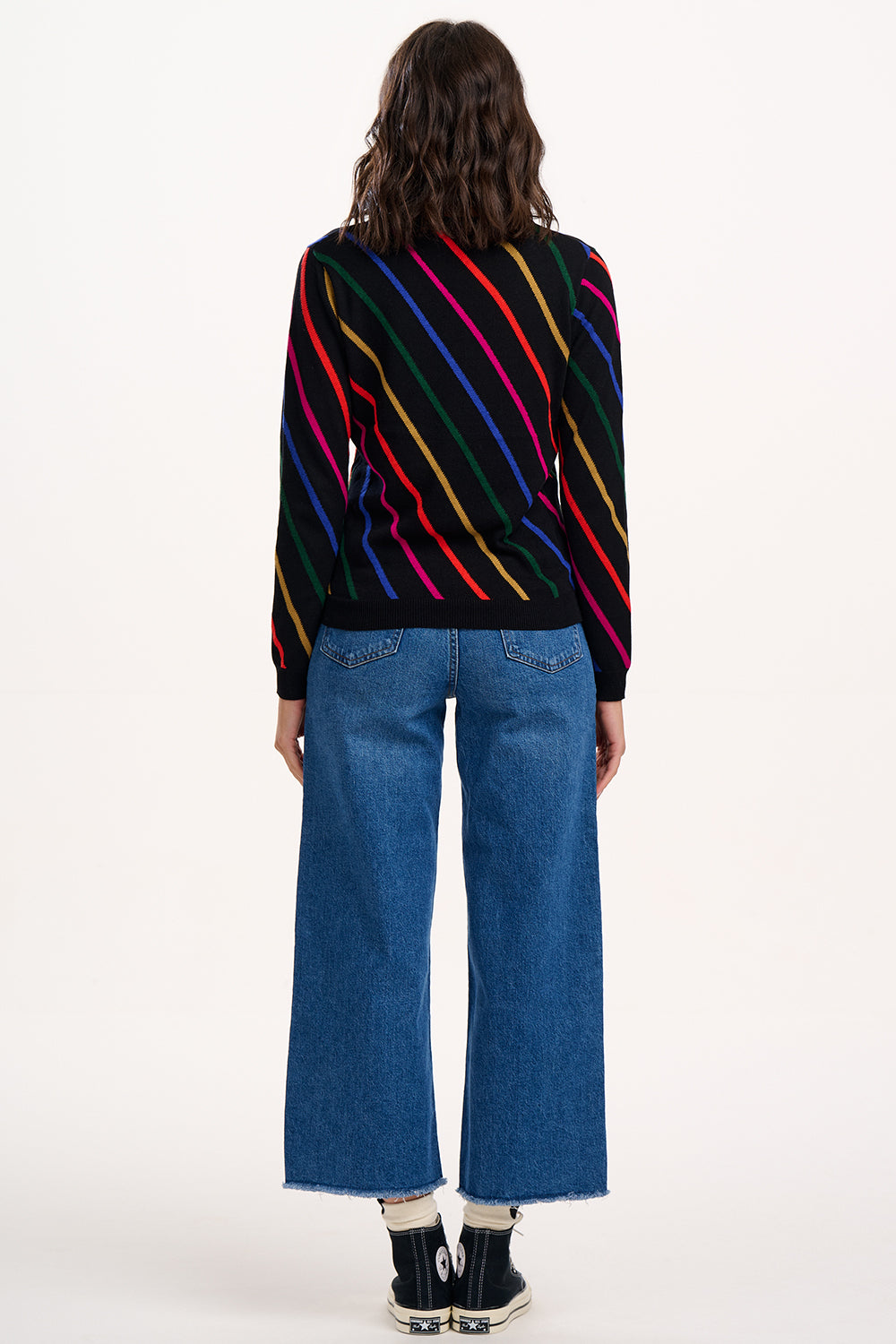 Person wearing a black sweater with colorful stripes and blue jeans on a white background