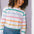 A woman wearing an off-white long-sleeve t-shirt with a thin rainbow stripe pattern over it, paired with blue jeans.