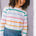 A woman wearing an off-white long-sleeve t-shirt with a thin rainbow stripe pattern over it, paired with blue jeans.