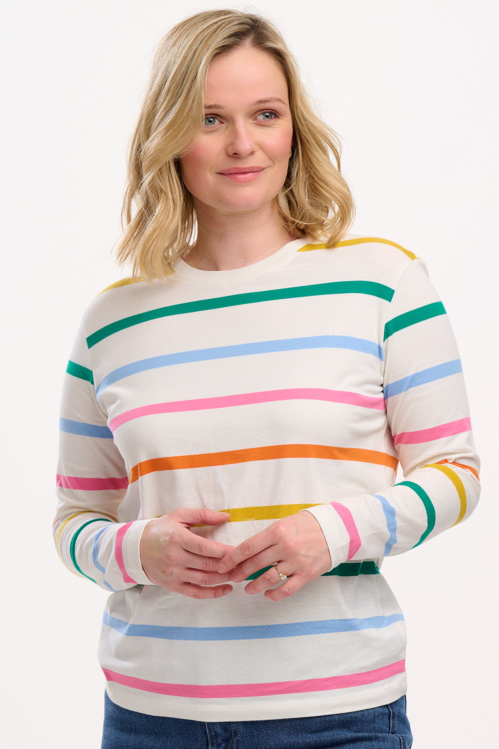 A woman wearing an off-white long-sleeve t-shirt with a thin rainbow stripe pattern over an off-white base, paired with jeans.