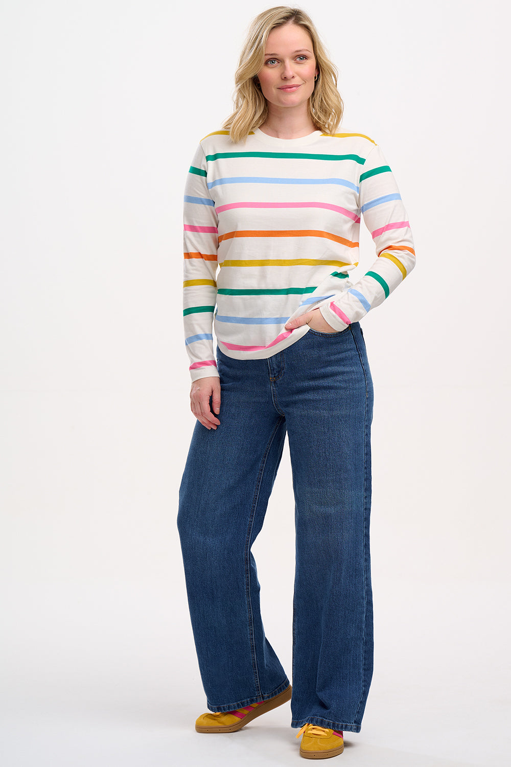 A woman wearing an off-white long-sleeve t-shirt with a thin rainbow stripe pattern over an off-white base, paired with jeans.