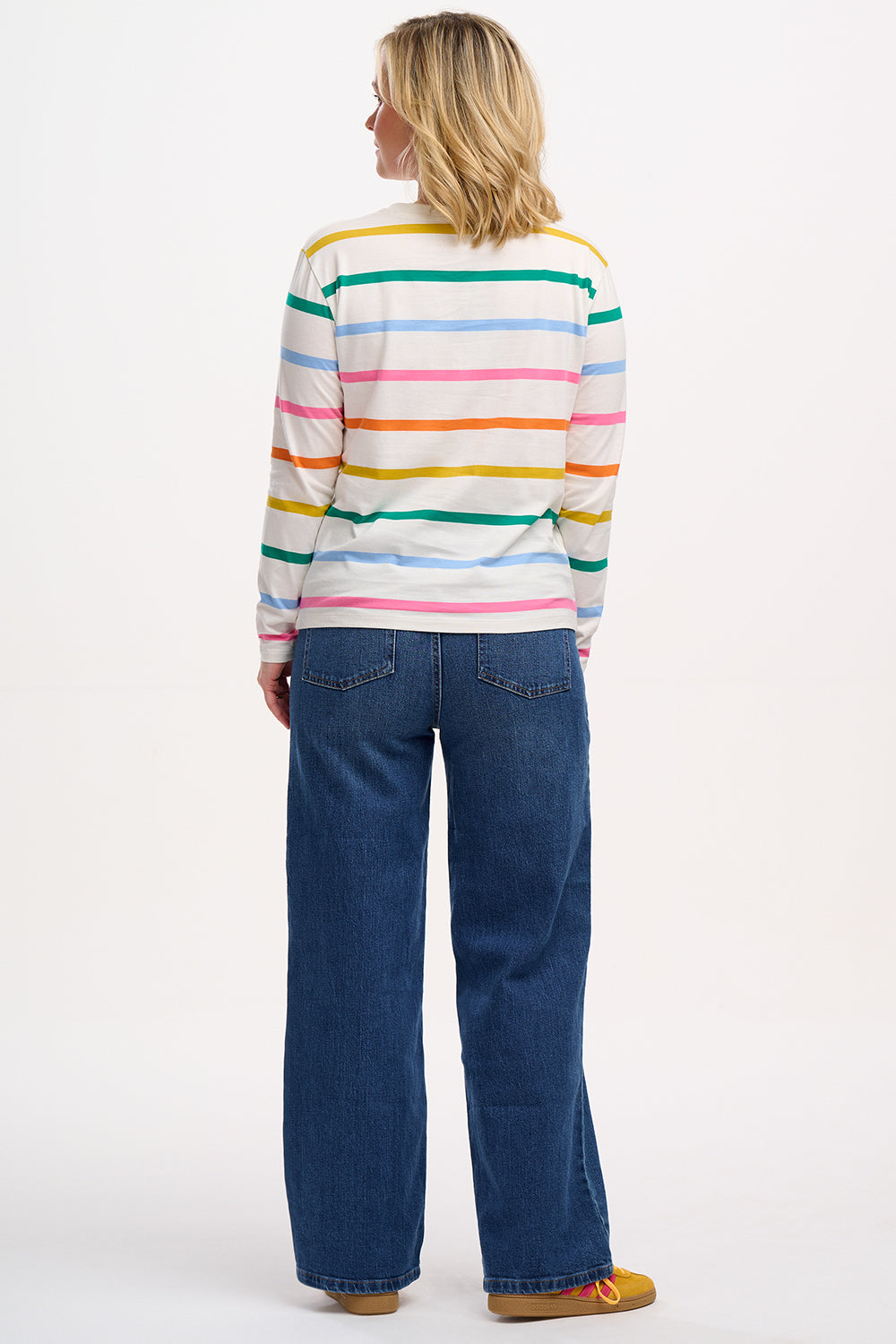 A woman wearing an off-white long-sleeve t-shirt with a thin rainbow stripe pattern over an off-white base, paired with jeans.
