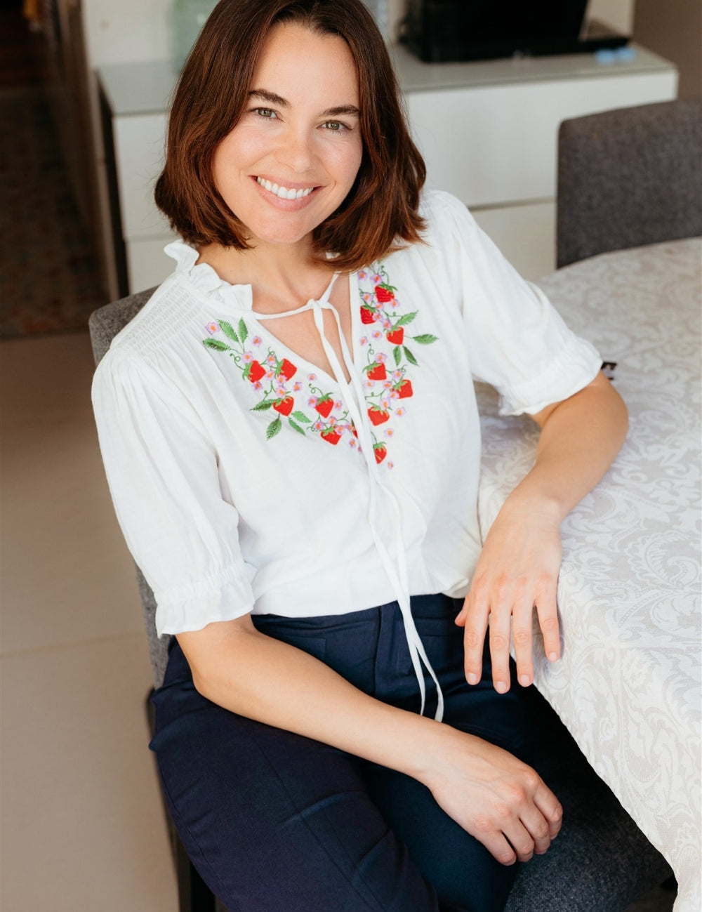 Woman wearing a white blouse with red floral embroidery sitting on a couch.