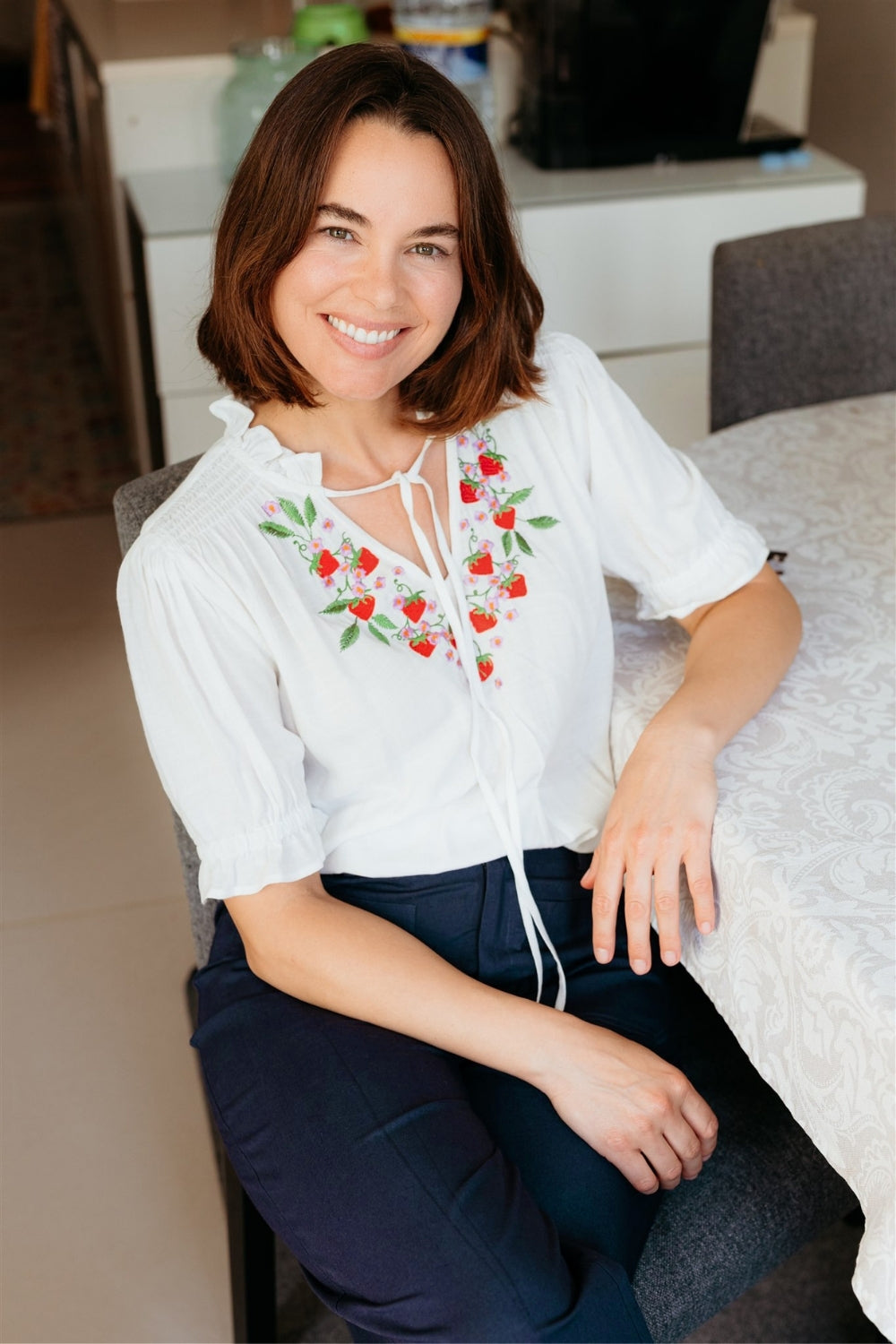 Woman wearing a white blouse with red floral embroidery sitting on a couch.
