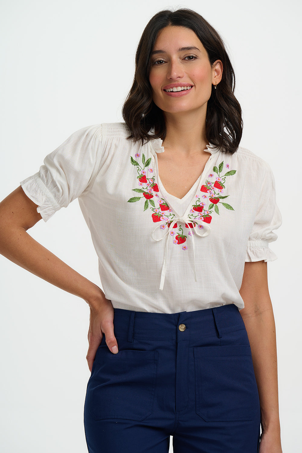 Woman wearing a white blouse with floral embroidery and navy pants on a white background