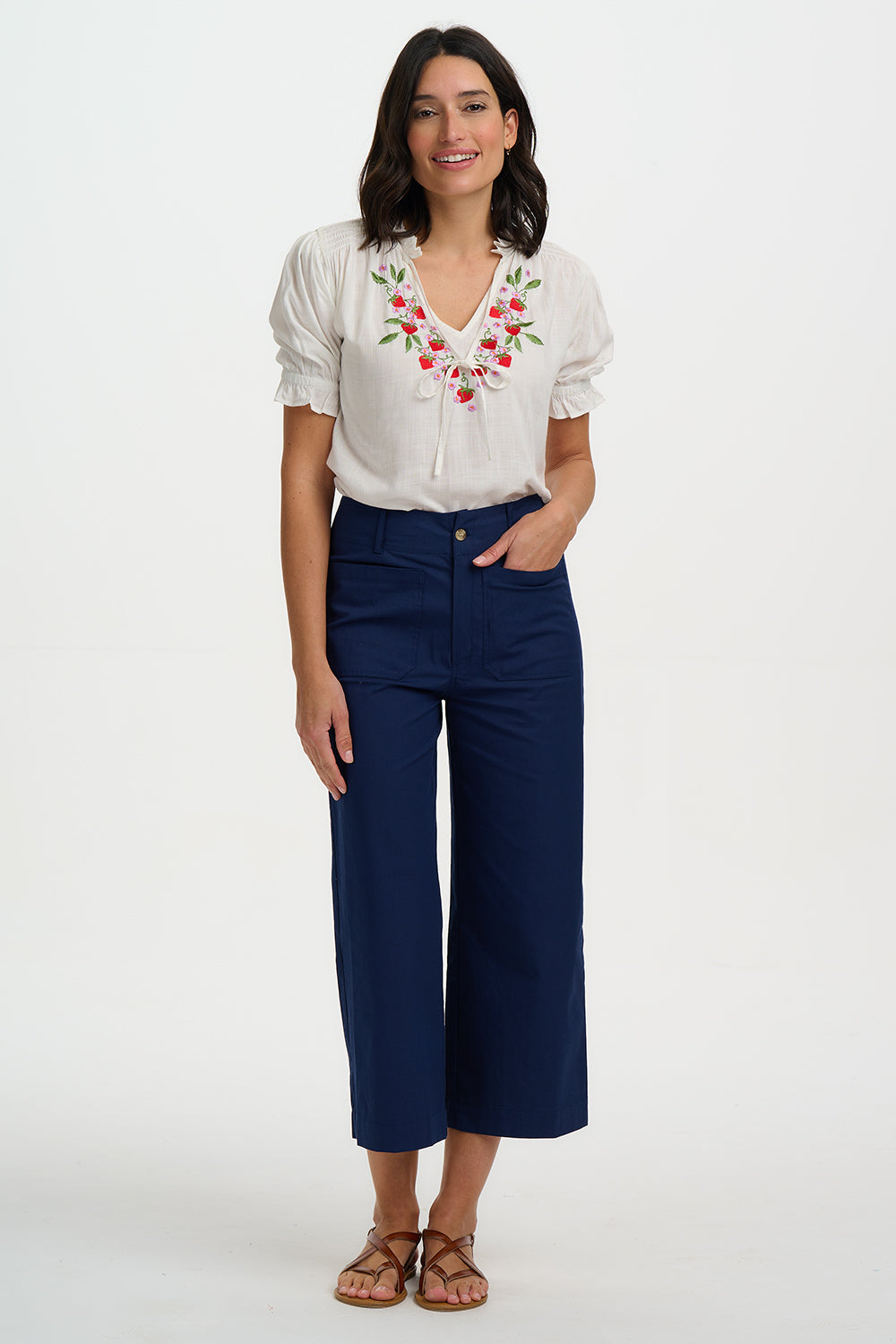 Woman wearing a white blouse with floral embroidery and navy pants on a white background