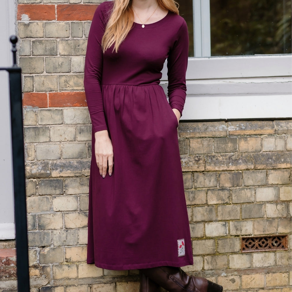 A woman wearing a long-sleeve, knee-length burgundy dress with side pockets, standing outdoors against a brick wall.