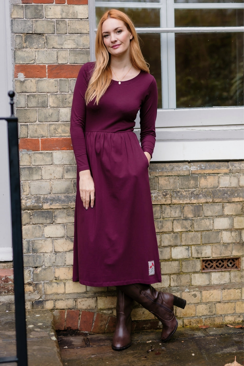 A woman wearing a long-sleeve, knee-length burgundy dress with side pockets, standing outdoors against a brick wall.