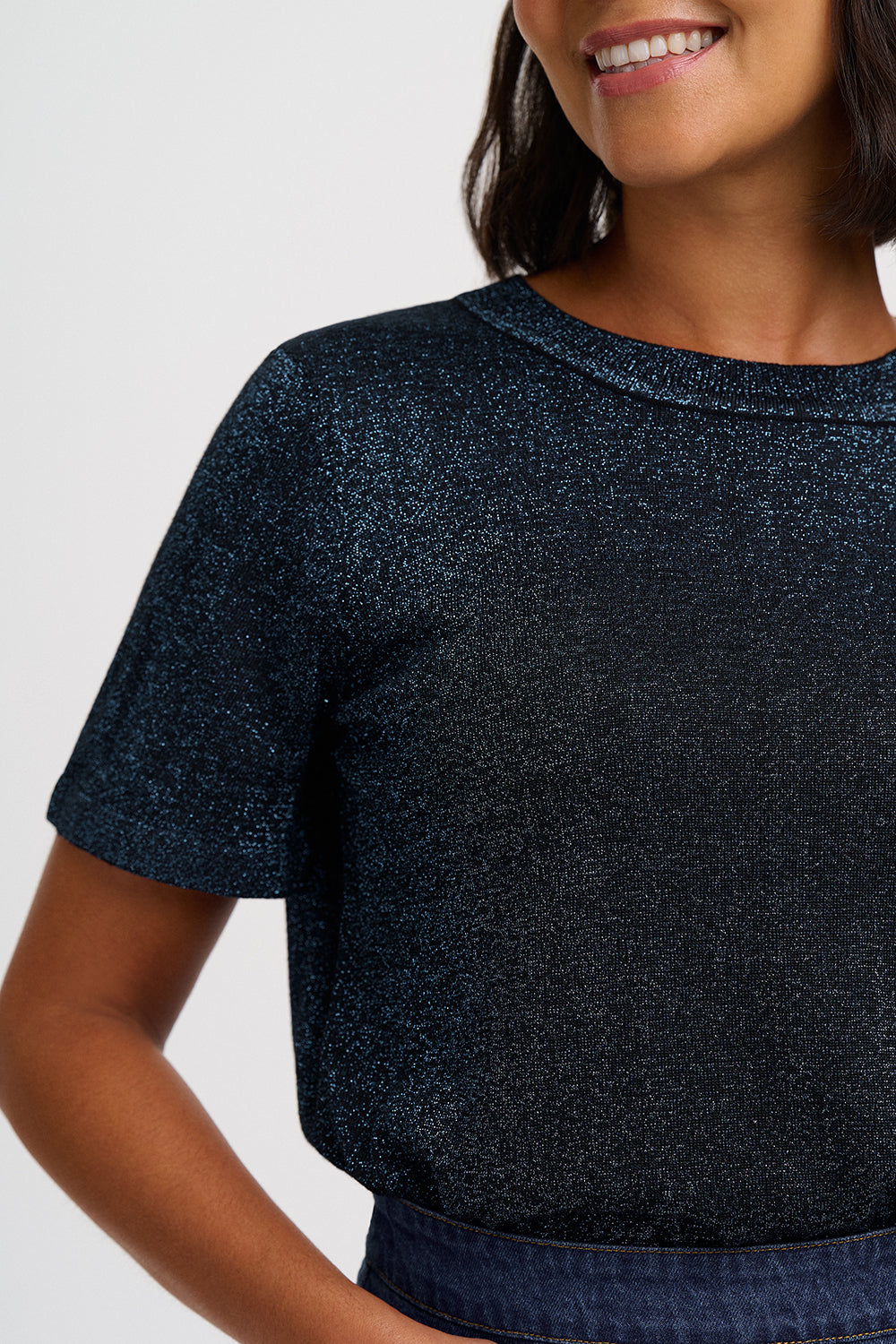 A close-up of a woman wearing a short-sleeve crewneck top with a dark navy blue glittery texture, paired with a denim skirt or shorts.