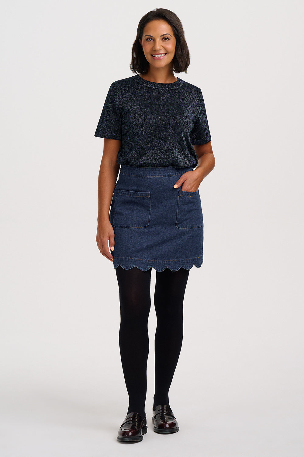 A woman modeling a dark navy blue short-sleeve knit top and a matching dark blue denim mini skirt with a scalloped hem, paired with black tights and dark brown leather loafers, against a plain light gray studio background.