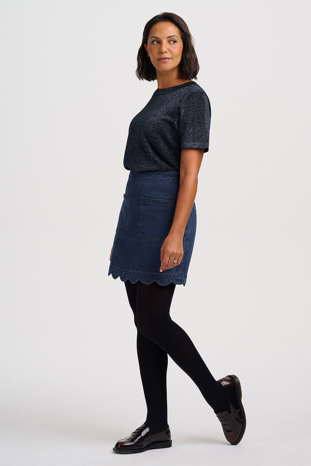 A woman modeling a dark blue textured short-sleeve top and a matching blue denim mini skirt with a scalloped hem, styled with black tights and black leather shoes against a light gray studio background.