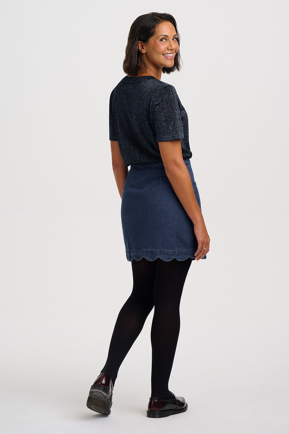 A woman modeling a dark navy blue short-sleeve knit top and matching denim mini skirt with a scalloped hem, paired with black tights and dark loafers, against a plain light gray studio background.