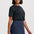 A woman modeling a dark charcoal gray short-sleeve knit top and a dark blue denim mini skirt with a scalloped hem and front patch pocket, against a light gray studio background.