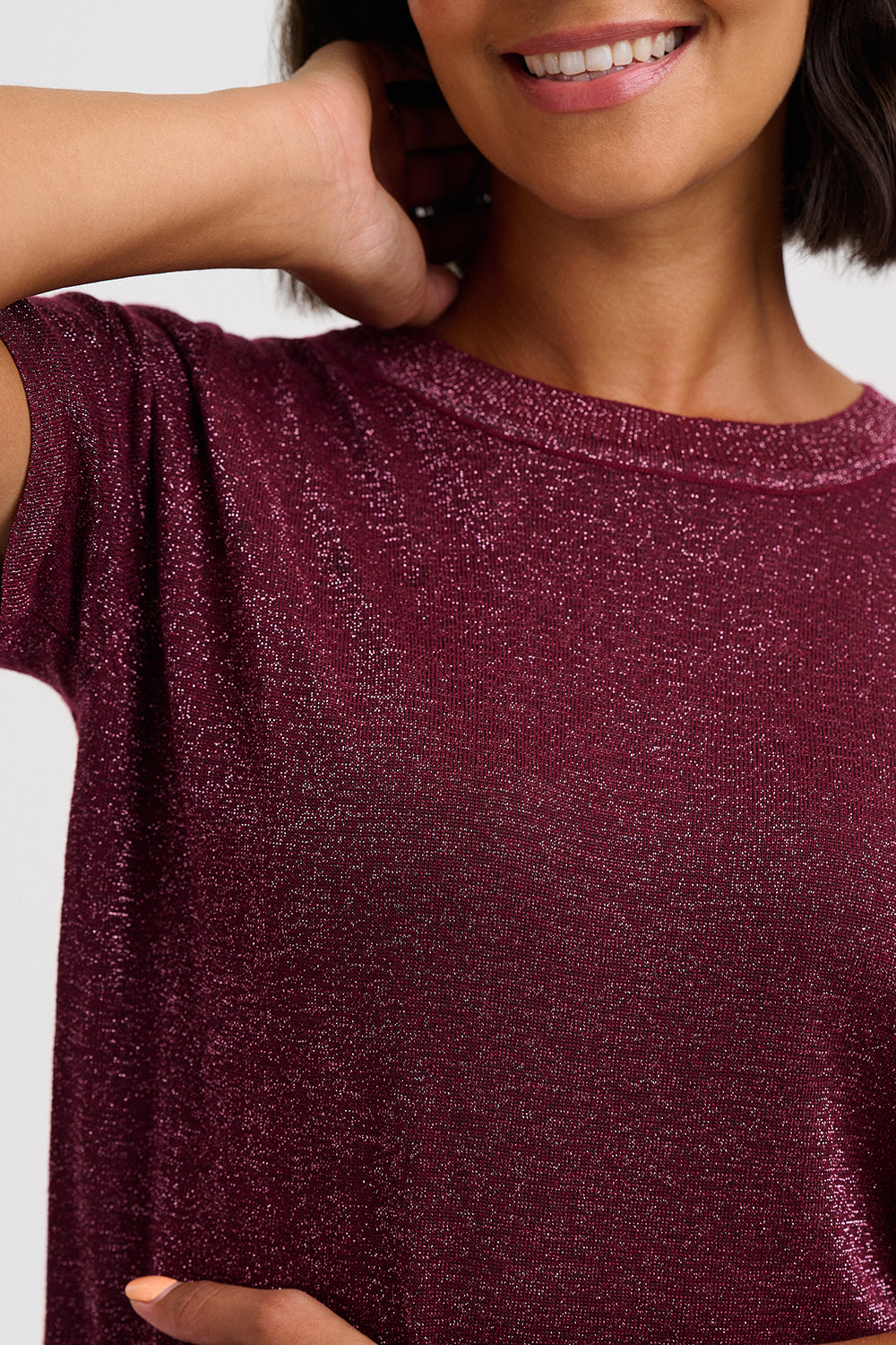 A close-up of a woman wearing a short-sleeve, crew-neck top made of sparkling, glittery burgundy fabric. The image is cropped to show her neck, lower face, and torso, with a plain light gray background.